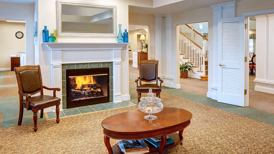 A cozy living room area with a lit fireplace surrounded by a white mantel and green tiles. Two wooden armchairs with leather seats are placed on either side of the fireplace. In front of the fireplace is an oval wooden coffee table with a glass jar on top. The room has beige patterned carpet and white walls with decorative columns. In the background, there is a staircase with wooden handrails and a partially open white door.