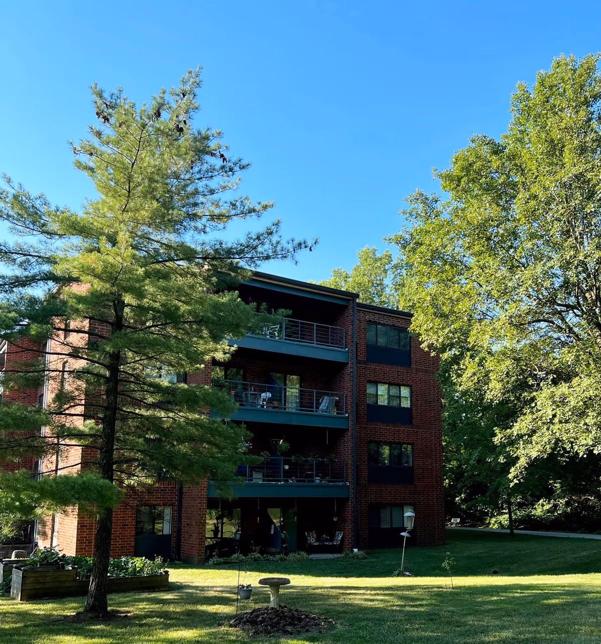 Three-story brick apartment building with balconies surrounded by trees and a grassy lawn under a blue sky.