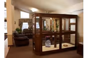 Interior view of a nursing home common area featuring a large wooden and glass enclosure in the center, surrounded by brown leather armchairs and windows with curtains in the background.