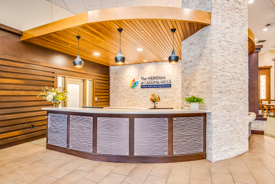Reception desk area inside The Meridian at Laguna Hills senior living community with a curved counter, modern pendant lights, a stone accent wall with the facility's logo, and decorative plants on the counter.