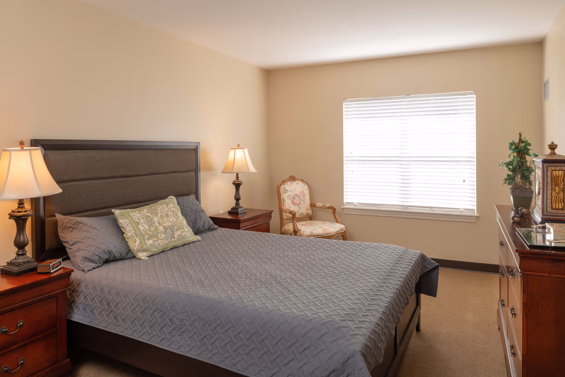 A bedroom with a large bed covered in a gray quilt and two pillows, flanked by two wooden nightstands each with a lamp. There is a decorative armchair near a window with closed blinds, and a wooden dresser with a plant and decorative items on top.