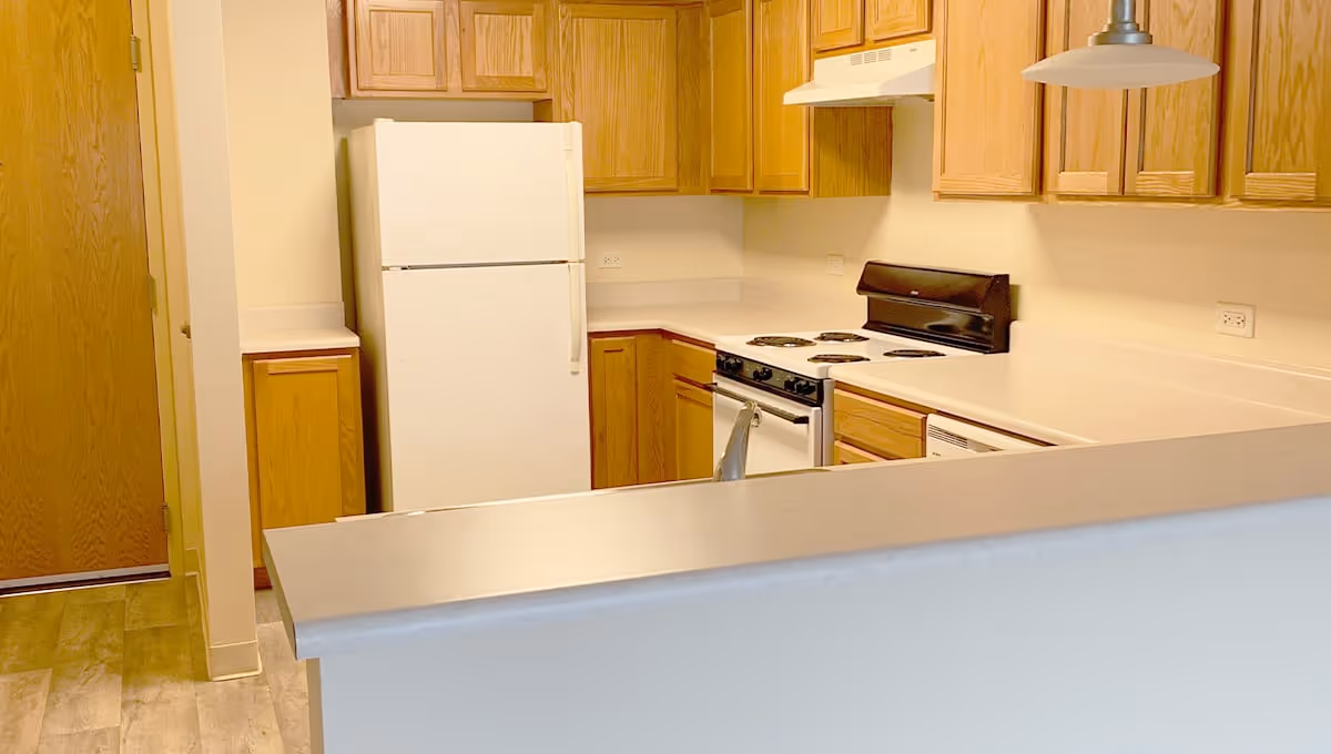 A kitchen area with wooden cabinets, a white refrigerator, a white stove with a black back panel, and a countertop with a sink. The floor has a wood-like finish and there is a hanging light fixture above the counter.