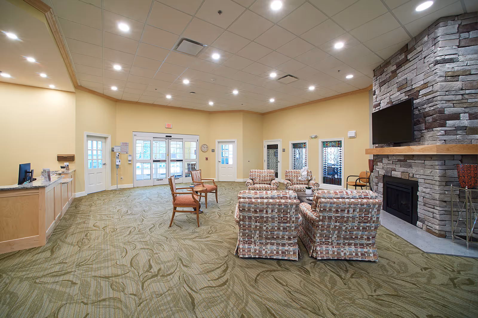 A spacious senior living facility common area with patterned armchairs arranged around a central coffee table, a stone fireplace with a mounted flat-screen TV, a reception desk on the left, and double glass doors leading outside. The room has beige walls, a green patterned carpet, and recessed ceiling lights.