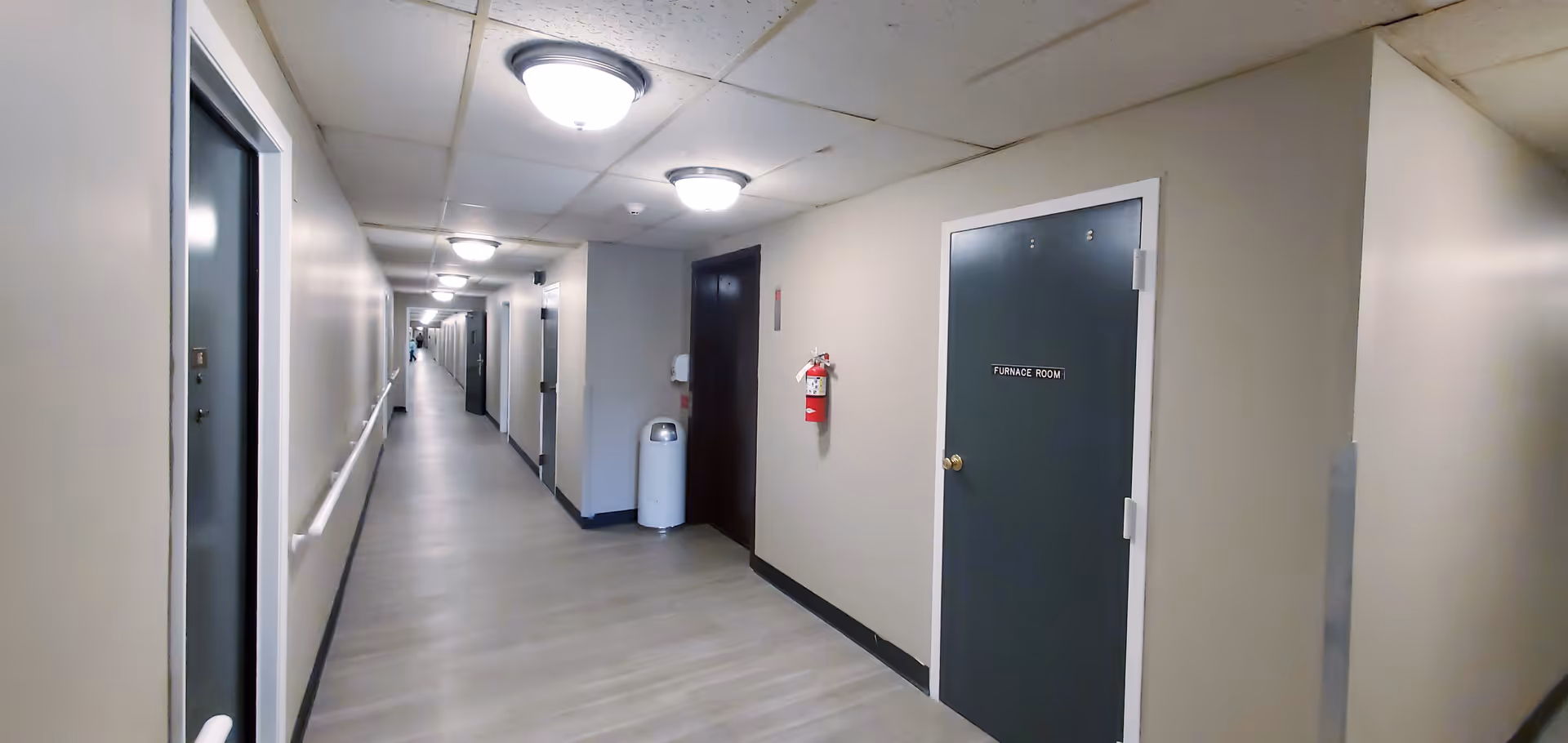 A long, well-lit hallway in Cougill Senior Apartments with beige walls, light wood flooring, and multiple doors on either side. The ceiling has several round light fixtures, and there is a fire extinguisher mounted on the wall next to a door labeled 'Furnace Room'. A trash can is also visible along the wall.