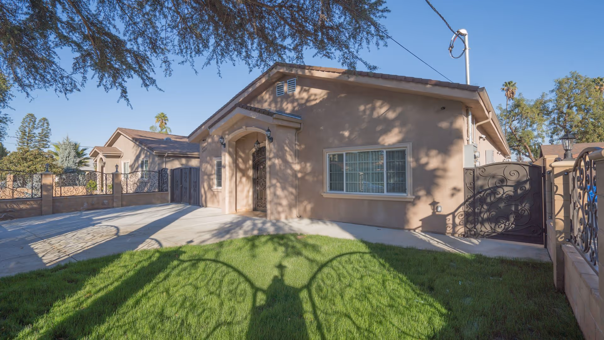 Front exterior of a single-story beige house with a gated driveway, small lawn, and decorative iron fence under a clear blue sky.