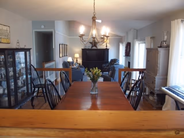 A cozy dining room with a wooden table and six black chairs. A vase with flowers is centered on the table. In the background, there is a living area with blue sofas, a fireplace, and a chandelier hanging above the dining table. The room has wooden floors and light-colored walls with windows letting in natural light.
