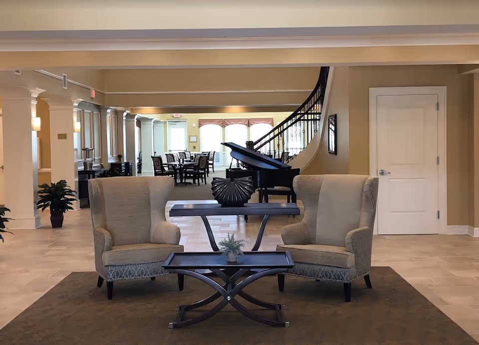 A senior living lobby/lounge with two armchairs and tables facing a grand piano, with a dining area and staircase in the background.