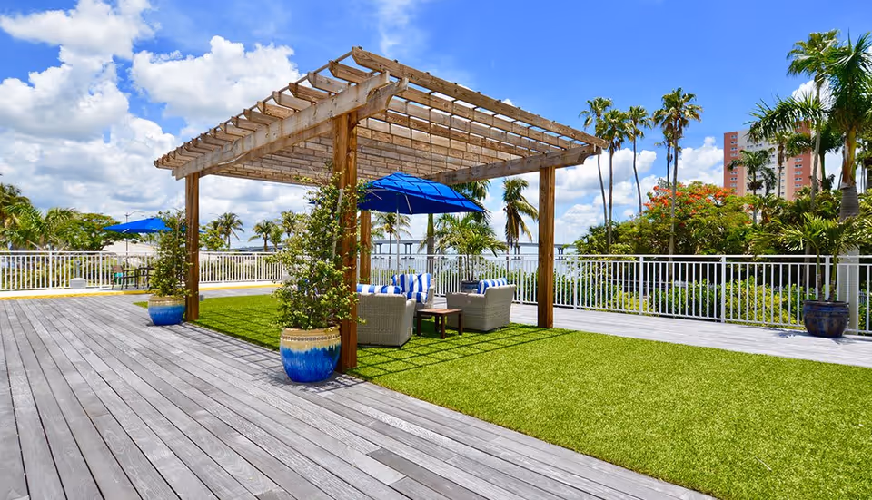 Outdoor patio area with a wooden pergola over a seating arrangement consisting of wicker chairs with blue and white striped cushions and a blue umbrella. The area has artificial grass and wooden decking, surrounded by potted plants and palm trees under a partly cloudy blue sky.