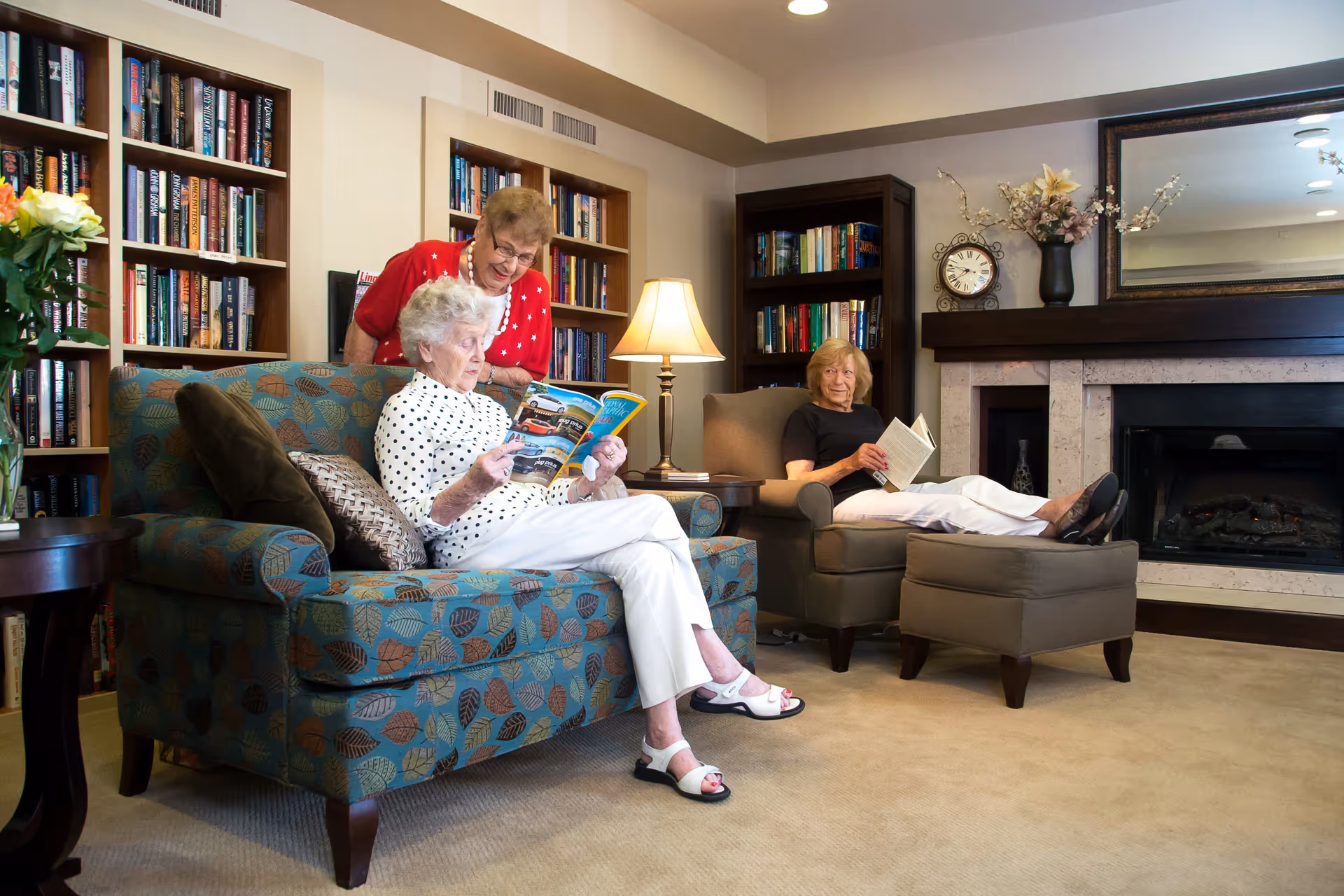Three elderly women sitting and reading in a comfortable library-style lounge with bookshelves and a fireplace.