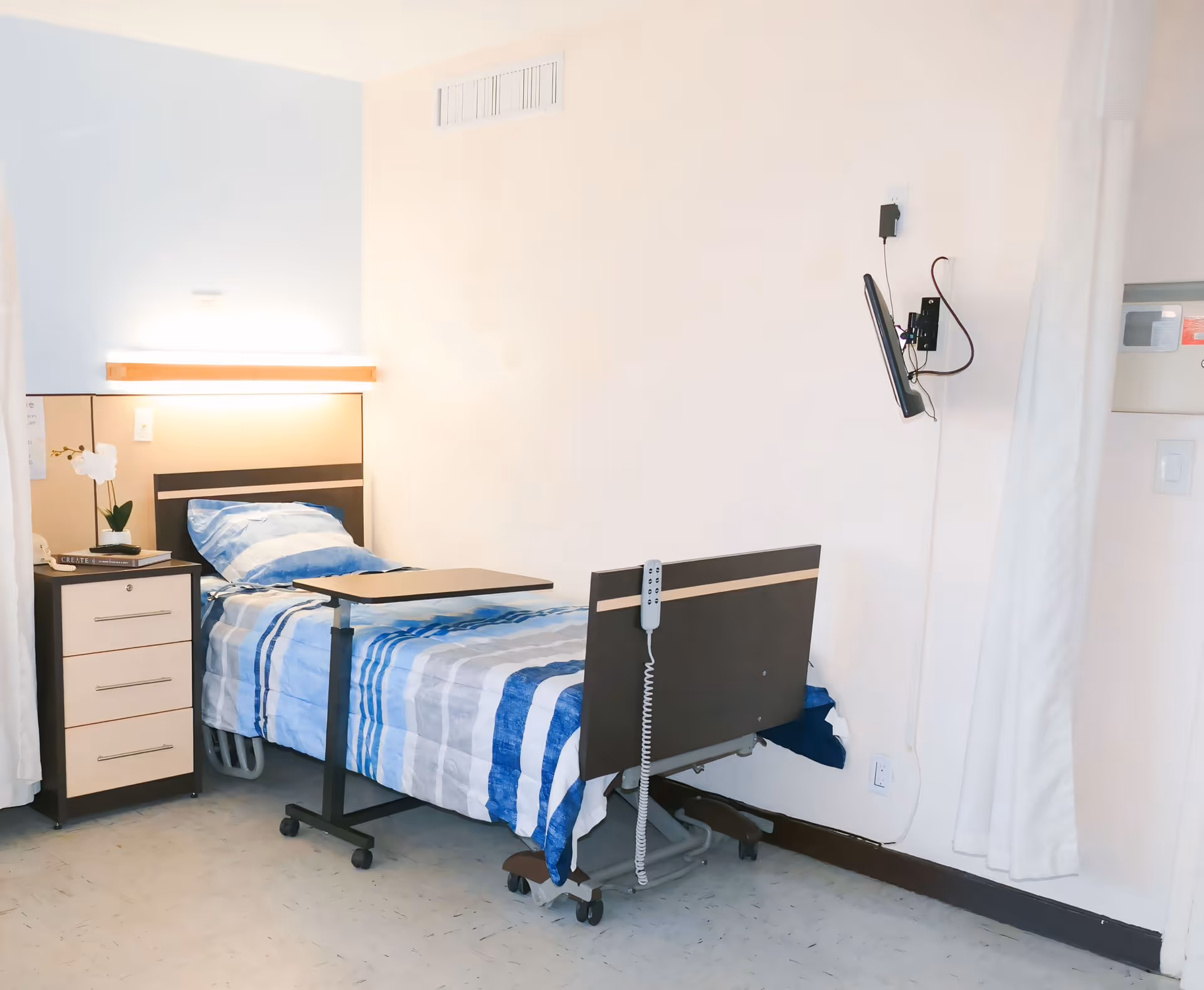 A clean and simple senior care facility bedroom with a single hospital-style bed covered in blue and white striped bedding. Next to the bed is a small bedside table with drawers, a white orchid plant, and some books. A wall-mounted TV is positioned in front of the bed, and a white curtain hangs to the right side of the room. The room has light-colored walls and a tiled floor.