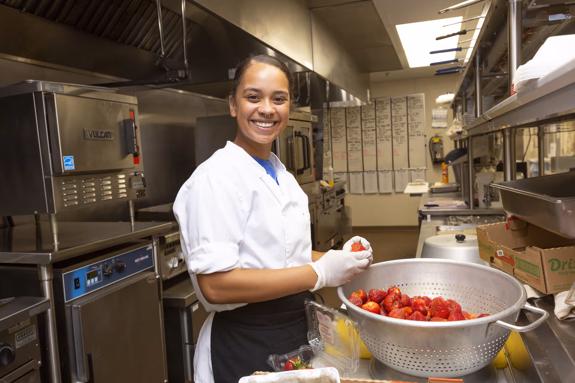 A smiling chef wearing a white coat and gloves is preparing strawberries in a commercial kitchen. She is standing next to a large metal colander filled with strawberries, with kitchen equipment and shelves visible in the background.