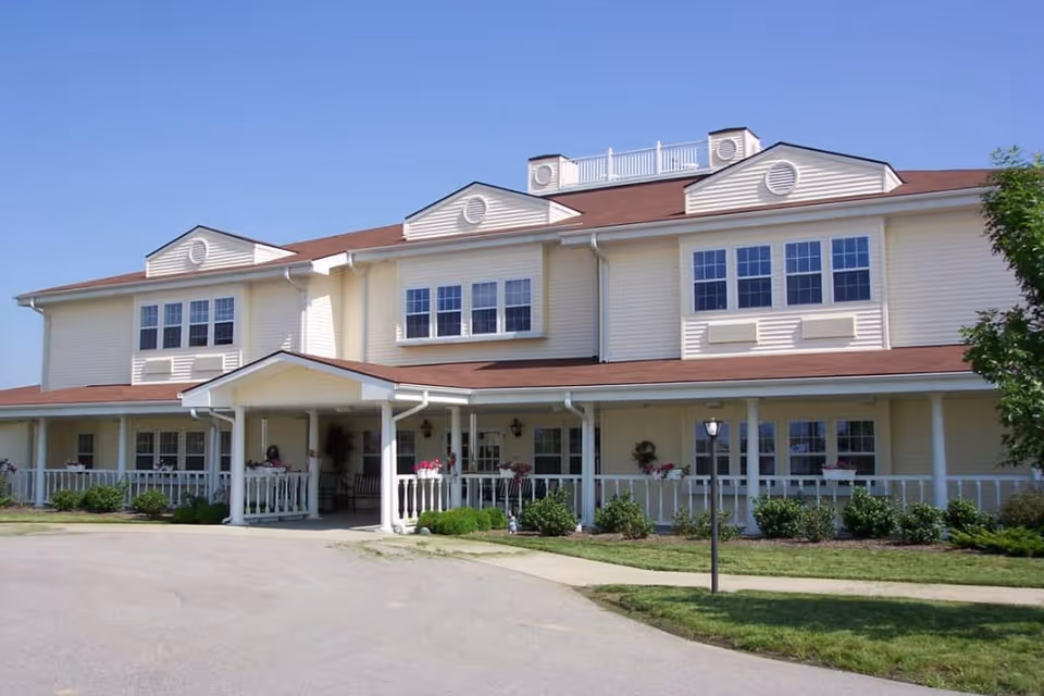 Exterior view of a two-story senior living facility building with beige siding and a red roof under a clear blue sky. The building has multiple windows, a covered entrance with white pillars, a white railing along the porch, and landscaped bushes and flowers in front.