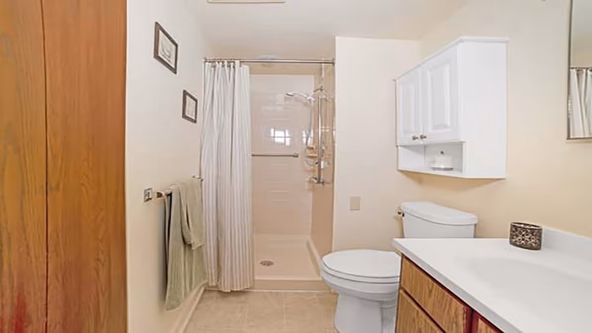 A clean bathroom featuring a walk-in shower with a white curtain, a toilet, a wooden vanity with a white countertop, a wall-mounted cabinet above the toilet, and two framed pictures on the wall. A towel hangs on a rack beside the shower.