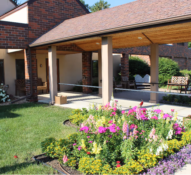 Covered entrance of a brick building with benches and a colorful flower bed in front.