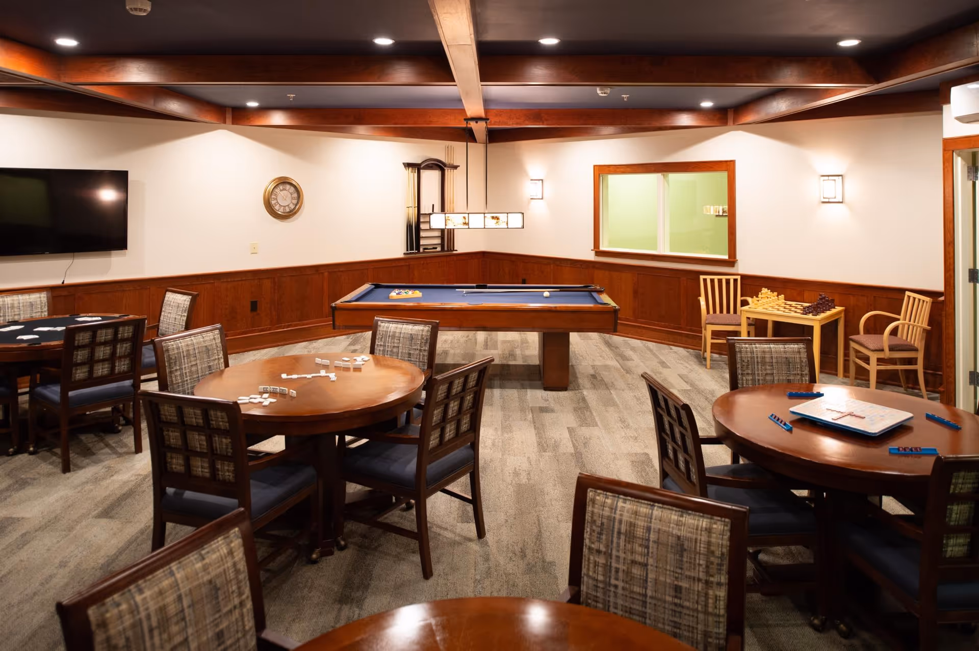 A well-lit game room with multiple round wooden tables and chairs arranged around them. The tables have various board games and tiles set up, including dominoes and Scrabble. In the center of the room is a pool table with a blue felt surface and pool cues. The walls are decorated with wood paneling and a clock, and there is a flat-screen TV mounted on one wall. The ceiling has wooden beams and recessed lighting.