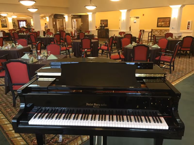 A black grand piano in the foreground with a dining area in the background featuring multiple tables covered with black tablecloths and set with folded green napkins and glassware. The dining chairs have red upholstery, and the room has warm yellow walls and carpeted floors.