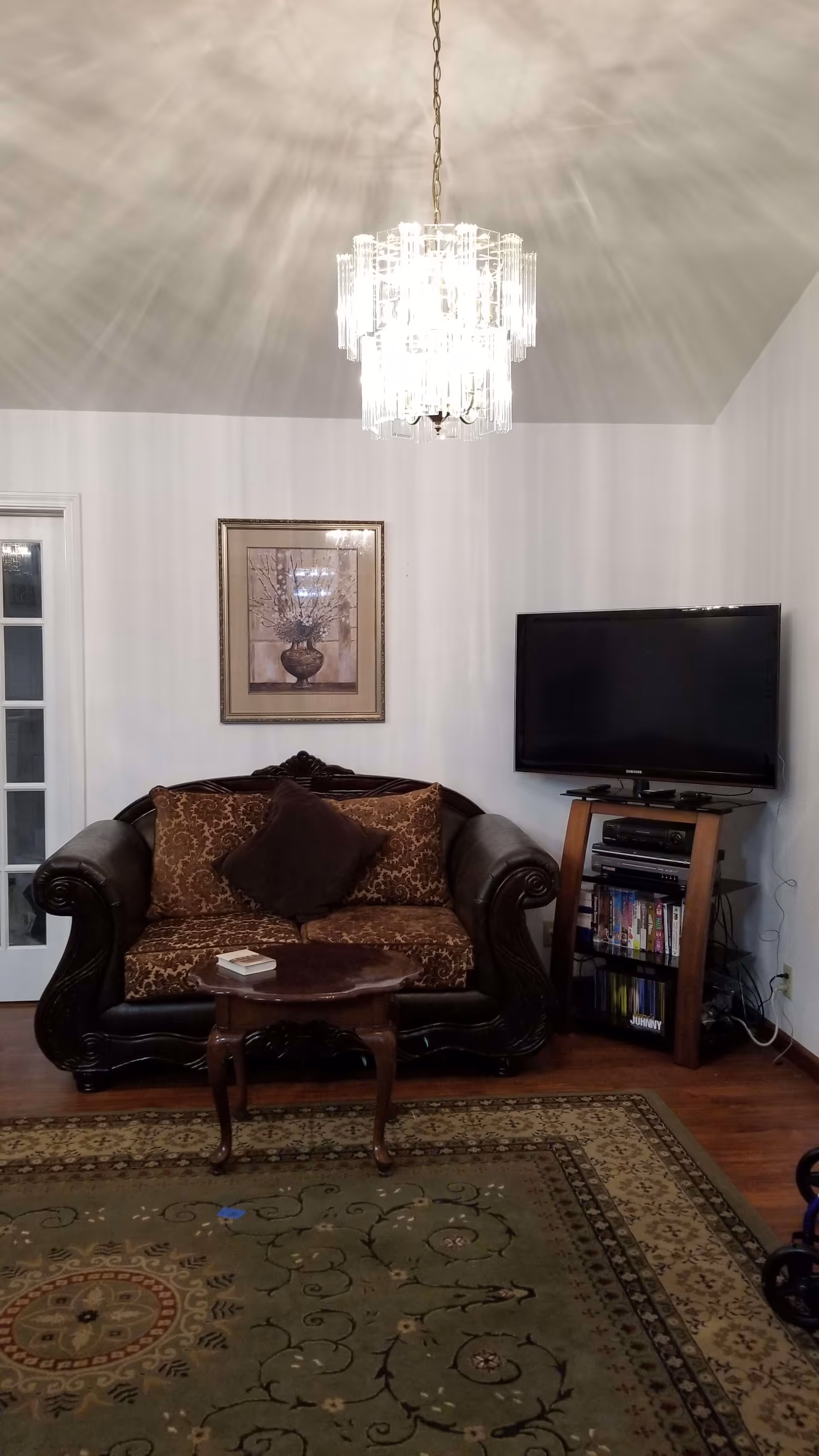 A cozy living room with a dark brown ornate loveseat with patterned cushions, a small wooden coffee table with a book on it, a flat-screen TV on a wooden stand with shelves holding DVDs and electronic devices, a framed floral artwork on the wall, and a decorative chandelier hanging from the ceiling. The floor has a patterned area rug and wooden flooring.