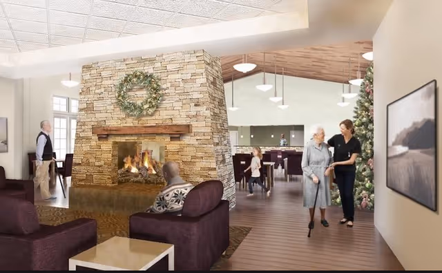 A cozy senior living common room with a central stone fireplace, seating area, dining space in the background, and residents with a staff member near a decorated Christmas tree.