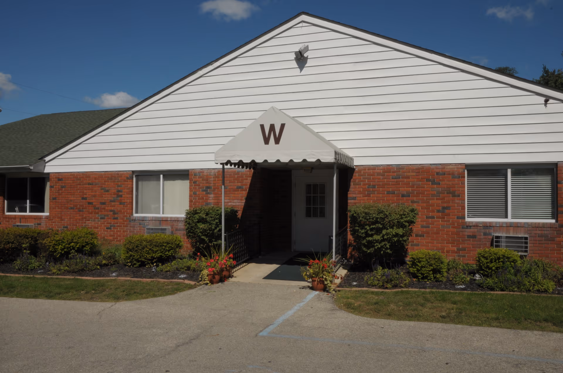 Exterior front view of a single-story building with red brick and white siding. The entrance has a small awning with the letter 'W' on it, surrounded by bushes and small plants. The sky is clear with a few clouds.