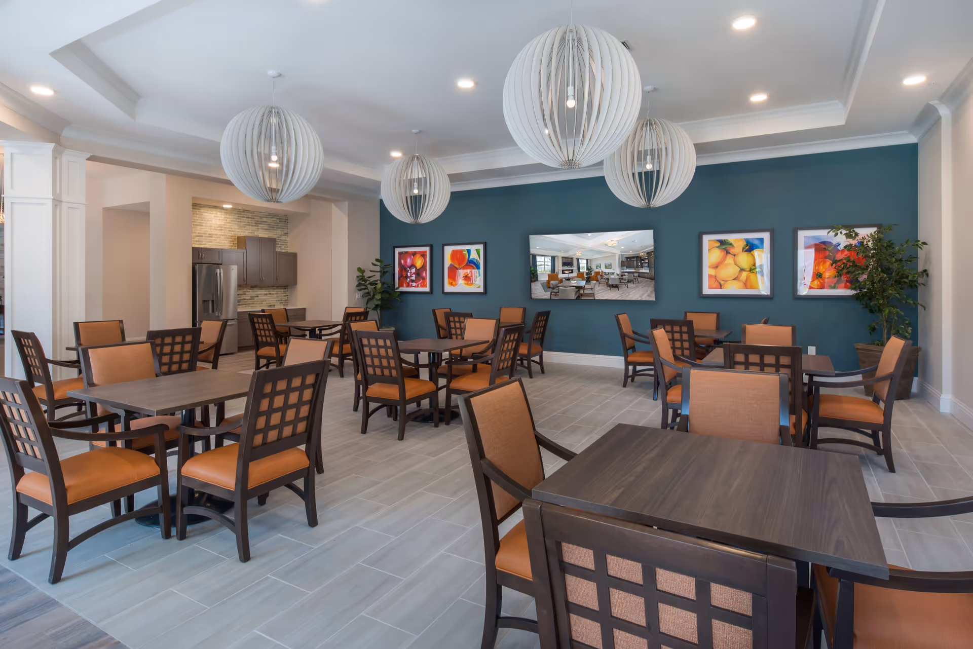 A spacious dining room with multiple dark wood tables and orange cushioned chairs. The room features a teal accent wall with colorful framed artwork and a large mirror. Three modern spherical pendant lights hang from the ceiling. In the background, there is a kitchen area with stainless steel appliances and a tiled backsplash. The floor is covered with light gray tiles.