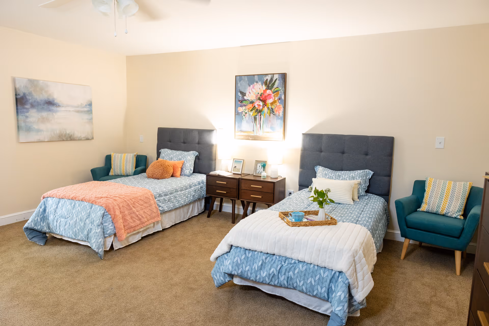 A bright, well-appointed room with two twin beds, a shared nightstand, wall art, and teal armchairs.