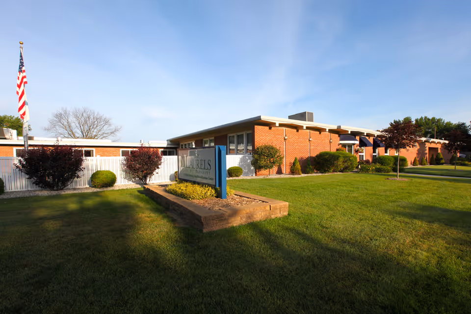 Exterior view of Laurels of Mt. Pleasant, a single-story brick building with a well-maintained lawn, shrubs, and small trees. An American flag is visible on the left side, and a sign with the facility's name is placed in the foreground.