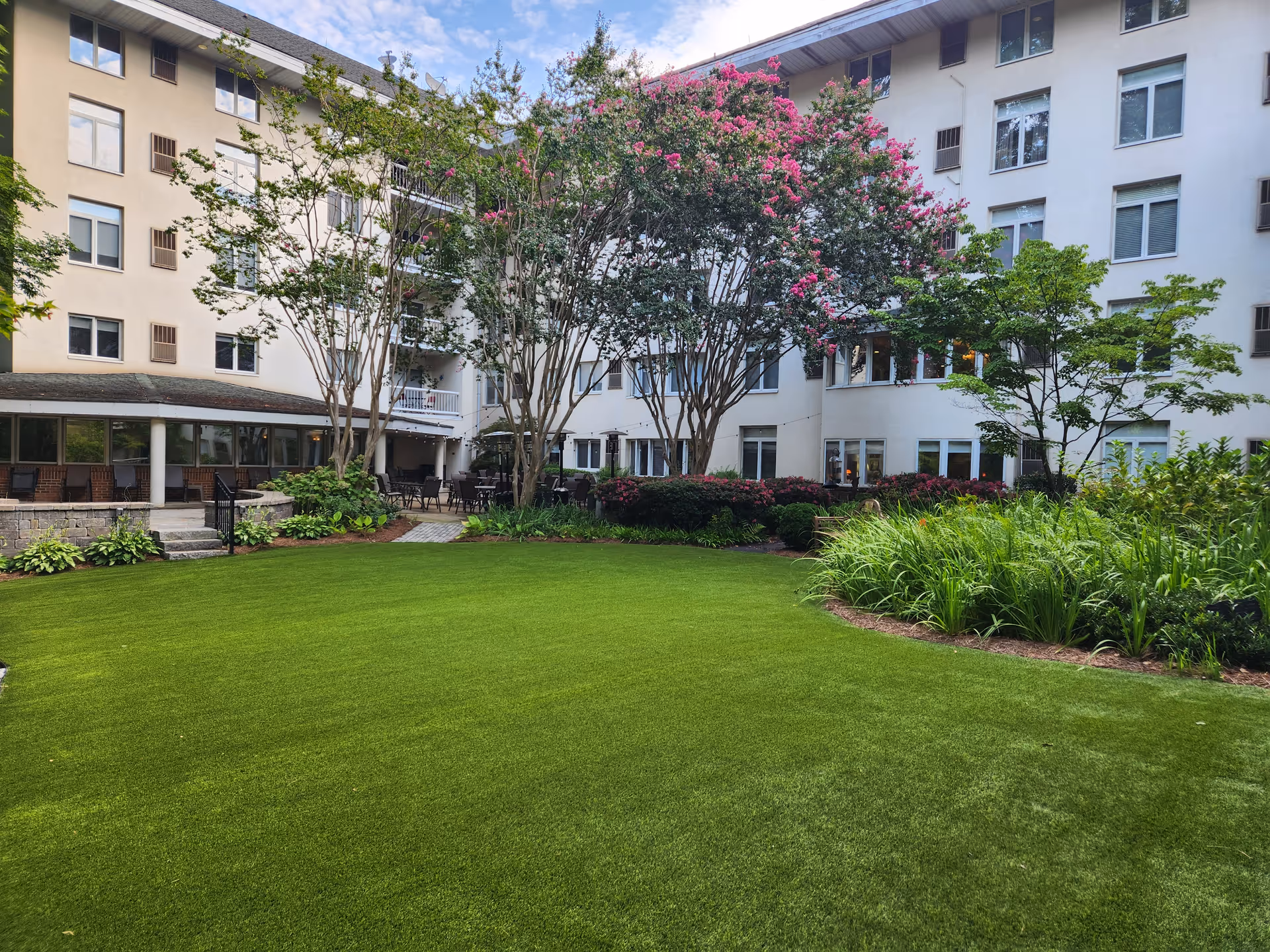 A well-maintained outdoor garden area at Saint Anne's Terrace with lush green grass, flowering trees with pink blossoms, various shrubs, and a multi-story building with many windows surrounding the garden.