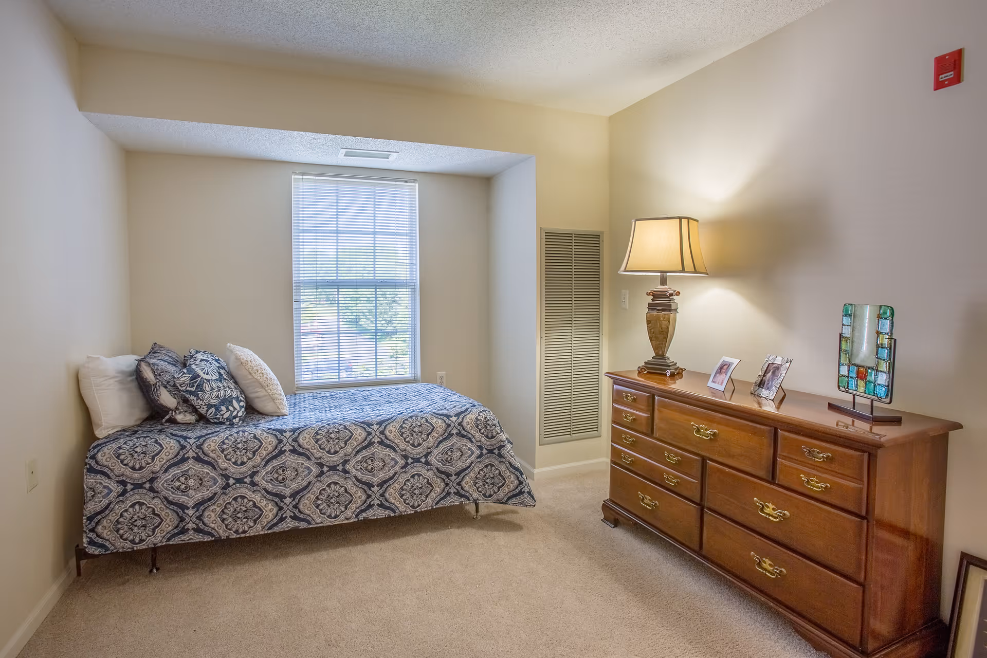 A small bedroom with a single bed covered in a blue and white patterned bedspread and several pillows. There is a window with blinds behind the bed. Opposite the bed is a wooden dresser with a lamp, two framed photos, and a decorative glass piece on top. The walls are light-colored and the floor is carpeted.