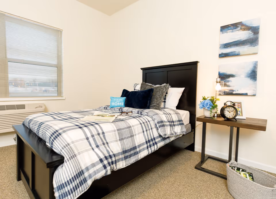 A neatly made single bed with plaid bedding in a bright bedroom, beside a nightstand with a lamp, clock and decor.