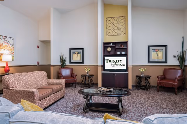 A cozy living room area in an assisted living facility featuring a patterned sofa, two red leather armchairs, a glass coffee table, side tables with small flower arrangements, framed artwork on the walls, and a TV cabinet displaying the Trinity Timbers logo.