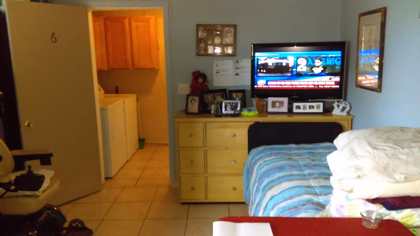 A small bedroom with a bed covered in a blue striped blanket and a pile of folded blankets on top. A dresser with multiple framed photos and a flat-screen TV mounted above it is against the wall. The TV is turned on displaying a news channel. To the left, an open door reveals a laundry area with a washing machine and wooden cabinets. The floor is tiled and the walls are painted light blue.