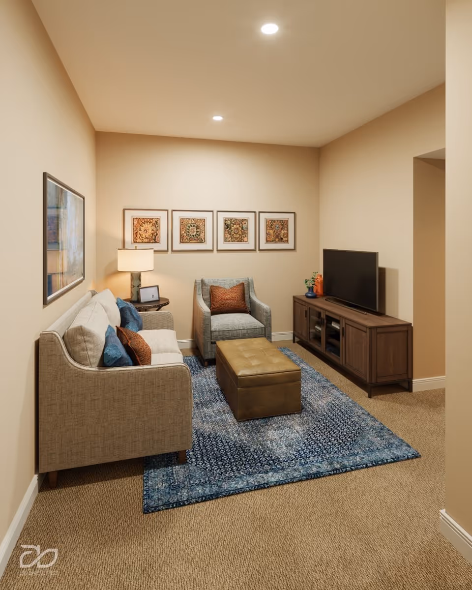 A cozy living room with beige walls and carpet, featuring a beige sofa with blue and brown cushions, a matching armchair with a brown cushion, a blue patterned area rug, a brown ottoman, a wooden TV stand with a flat-screen TV, a table lamp on a side table, and four framed artworks on the wall.