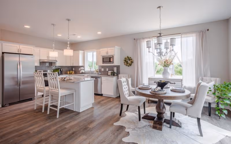 Bright and modern kitchen and dining area with white cabinetry, stainless steel appliances, a kitchen island with three white bar stools, a round wooden dining table set with four upholstered chairs, a large window with white curtains, and a light fixture above the table. The floor is wood with a light-colored rug under the dining table.