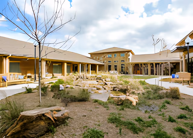 Outdoor courtyard area of Woodlands Place Rehabilitation Suites featuring a landscaped garden with pathways, benches, and surrounding single-story buildings under a partly cloudy sky.