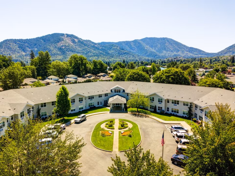 Aerial view of The Suites Assisted Living and Memory Care facility showing a large U-shaped building with a central entrance, surrounded by green lawns, trees, and parked cars. In the background, there are forested hills and a clear blue sky.