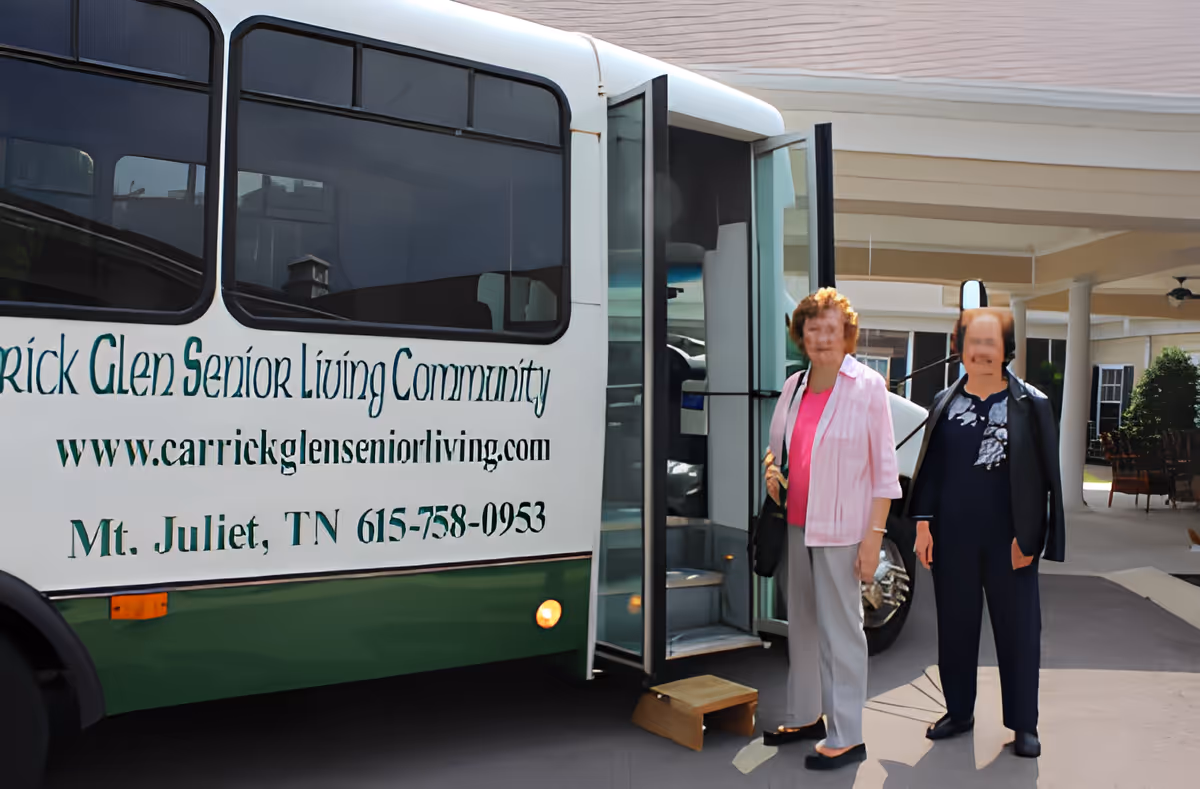 Two elderly women standing outside near the open door of a white and green shuttle bus with the text 'Carrick Glen Senior Living Community' and contact information on its side, in front of a covered entrance area of a building.