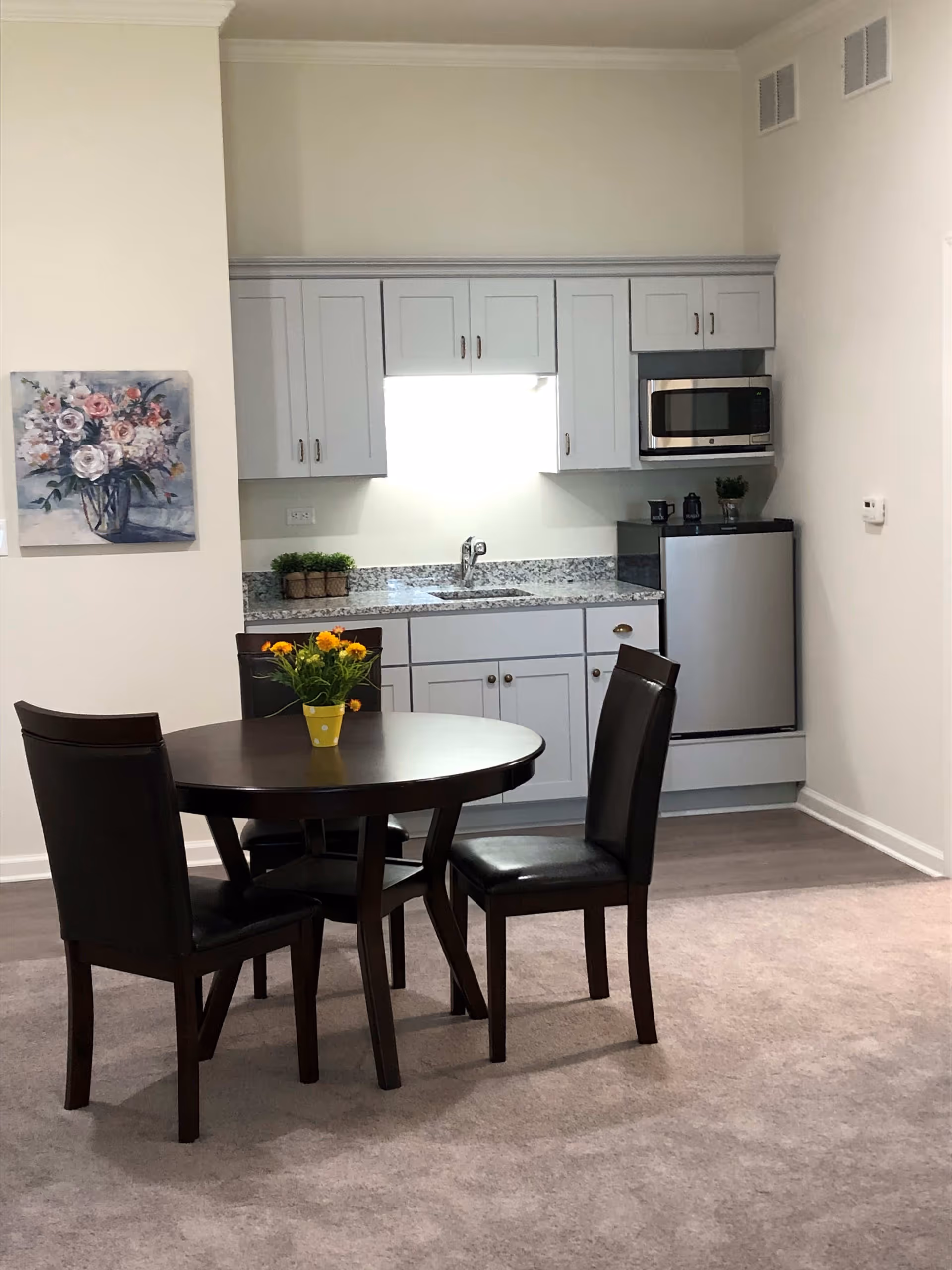 A small kitchenette area with light gray cabinets, a granite countertop, a stainless steel microwave, and a mini refrigerator. In front of the kitchenette is a round dark wooden dining table with four matching chairs. A small yellow pot with orange flowers is placed on the table. A floral painting hangs on the wall to the left.