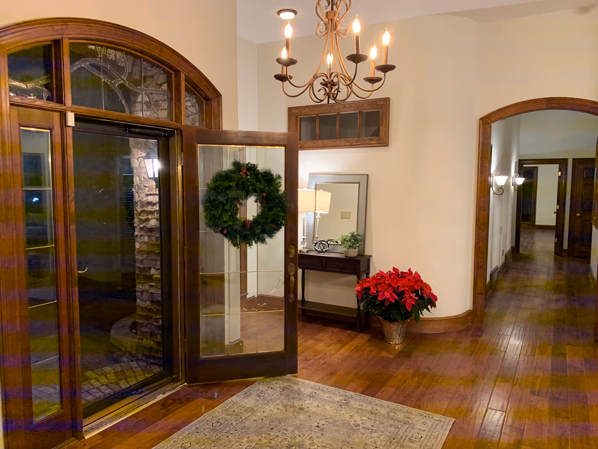 An entryway with a wooden floor and a glass door decorated with a green wreath. There is a small table with a lamp, a mirror, and a plant on it, as well as a poinsettia plant in a pot on the floor. A chandelier with lit bulbs hangs from the ceiling, and a hallway with wooden door frames is visible to the right.