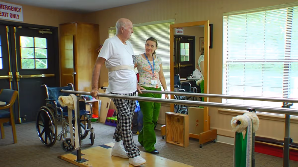 An elderly man wearing a white shirt and checkered pants is walking with the assistance of parallel bars in a rehabilitation or therapy room. A female caregiver or therapist in green pants and a patterned shirt is standing beside him, providing support and encouragement. There is a wheelchair and chairs in the background near a door with an emergency exit sign, and a large window with blinds letting in natural light.