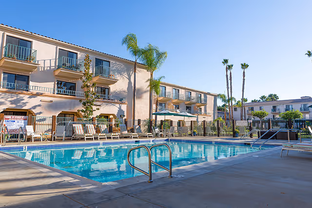 Outdoor swimming pool area at a senior living facility with lounge chairs, umbrellas, palm trees, and a three-story building in the background under a clear blue sky.