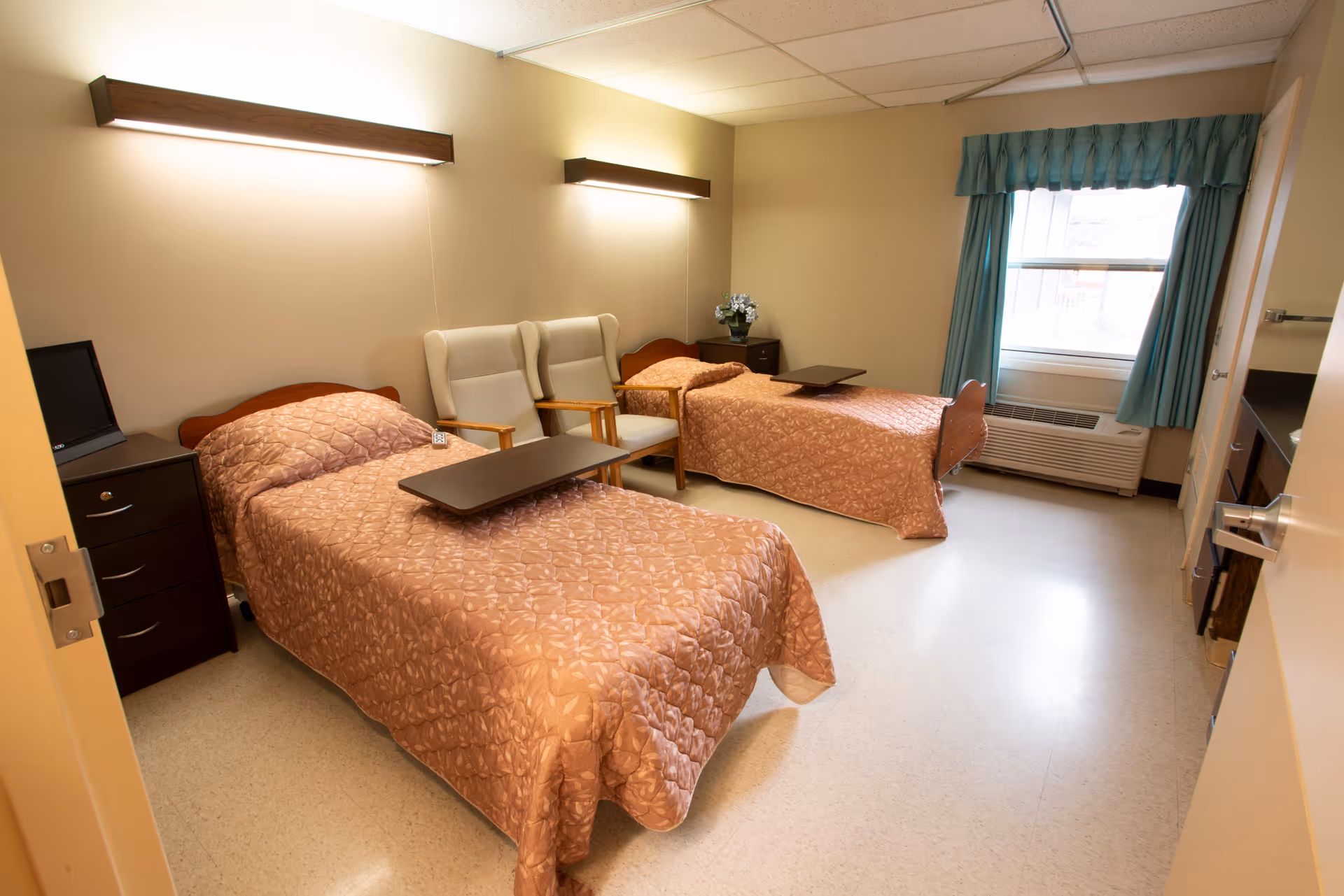 A shared bedroom in a senior living facility with two single beds covered in matching brown quilted bedspreads. Each bed has a dark brown overbed table. Between the beds are two beige armchairs with wooden armrests. The room has beige walls, a window with teal curtains, a dark brown nightstand with a small flower arrangement, and a heating/cooling unit below the window. The floor is light-colored and the lighting is provided by two wall-mounted fixtures above the beds.