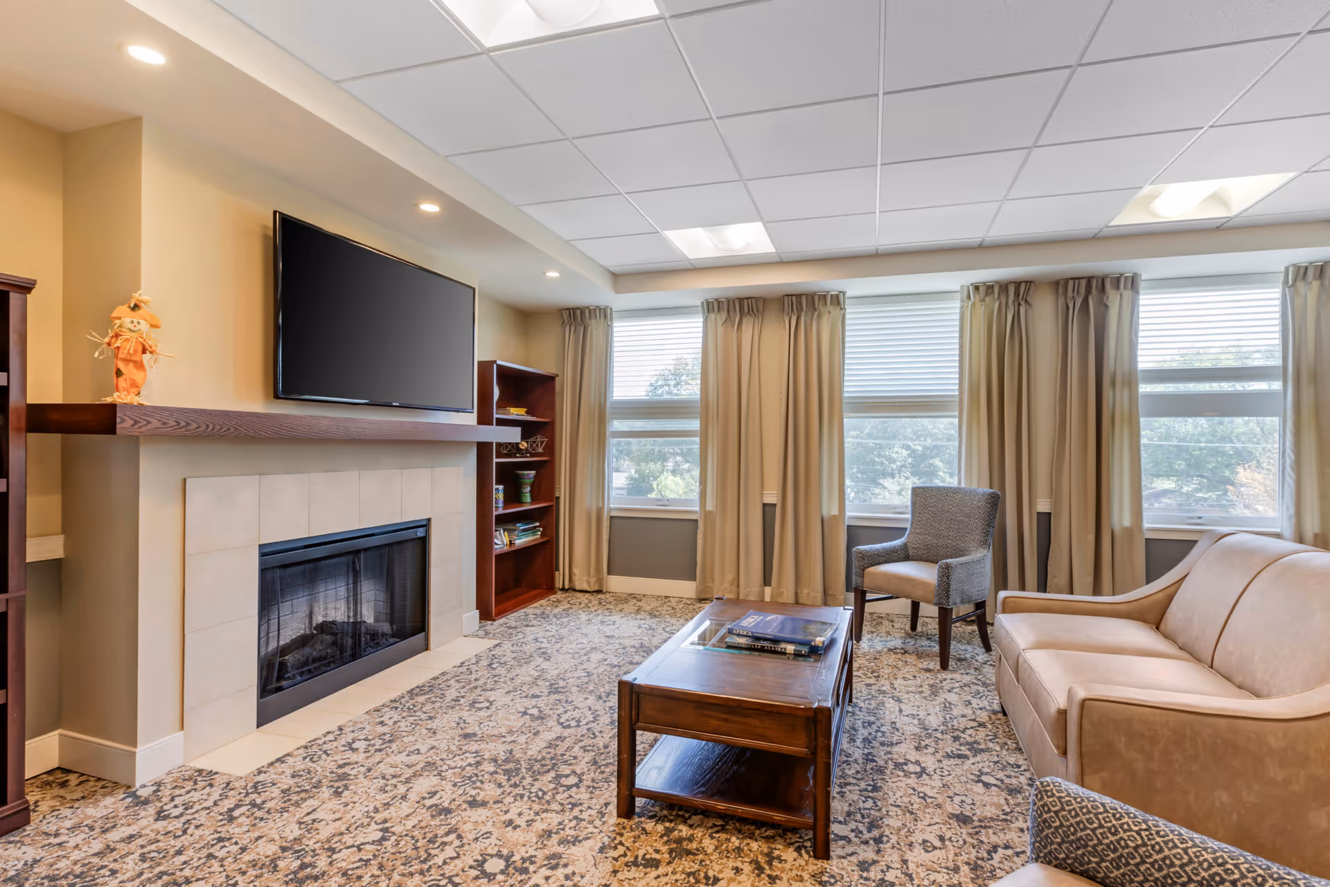 A cozy living room with a beige sofa, a gray armchair, a wooden coffee table with books on it, a fireplace with a mounted flat-screen TV above it, and large windows with beige curtains letting in natural light.
