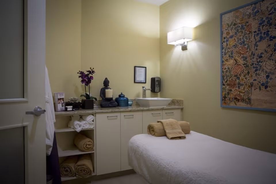 A serene massage or therapy room with a massage table covered in a white sheet and beige towels neatly rolled on top. The room has soft yellow walls, a countertop with decorative items including a purple orchid, a Buddha statue, candles, and a blue jar. There is a modern sink with a soap dispenser mounted on the wall above it. A framed floral artwork hangs on the right wall, and a wall-mounted light fixture provides soft illumination.