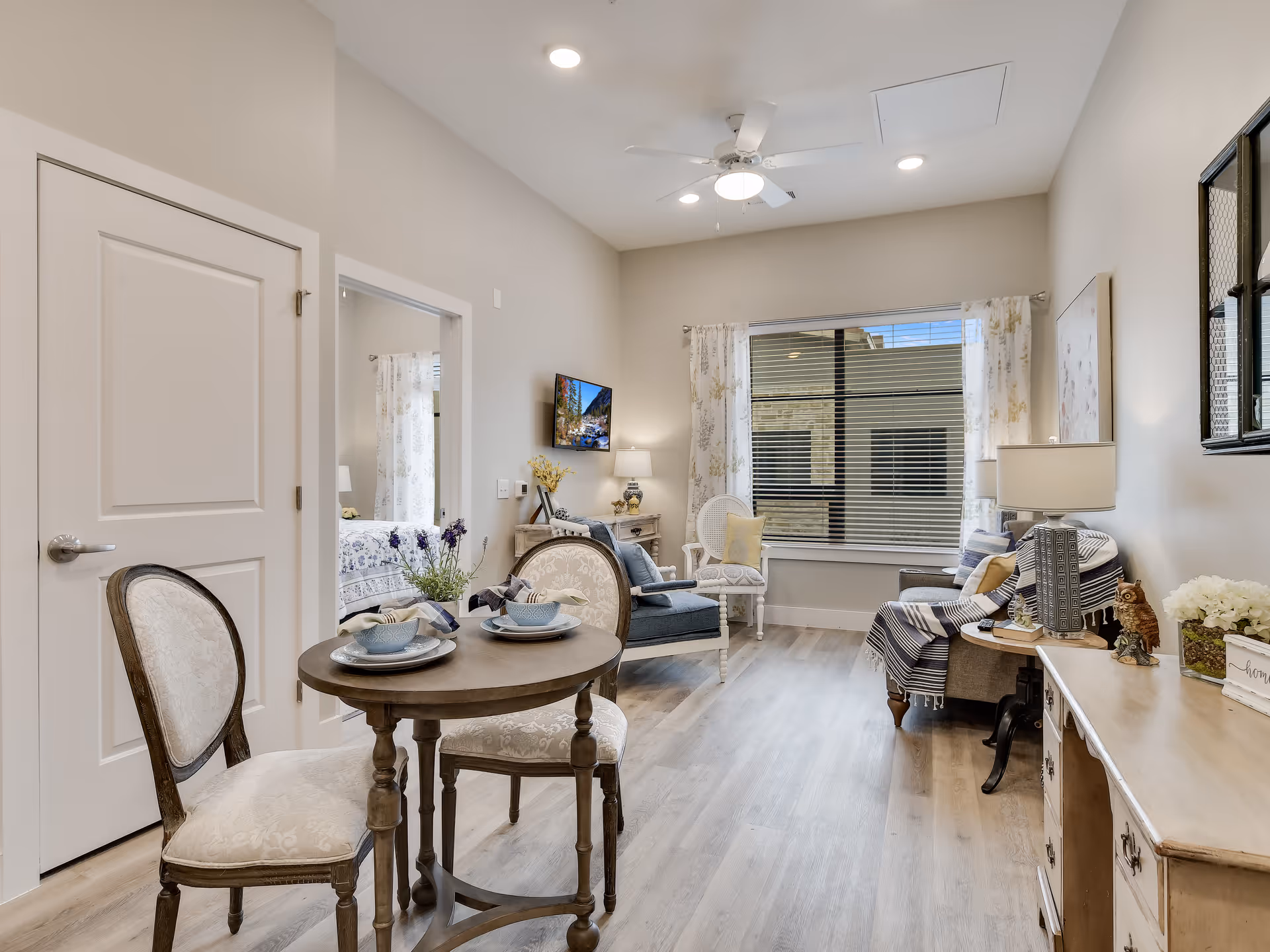 Well-lit living room and dining area with a round table and two chairs in the foreground, a sofa and armchair by a large window, and a wall-mounted TV.