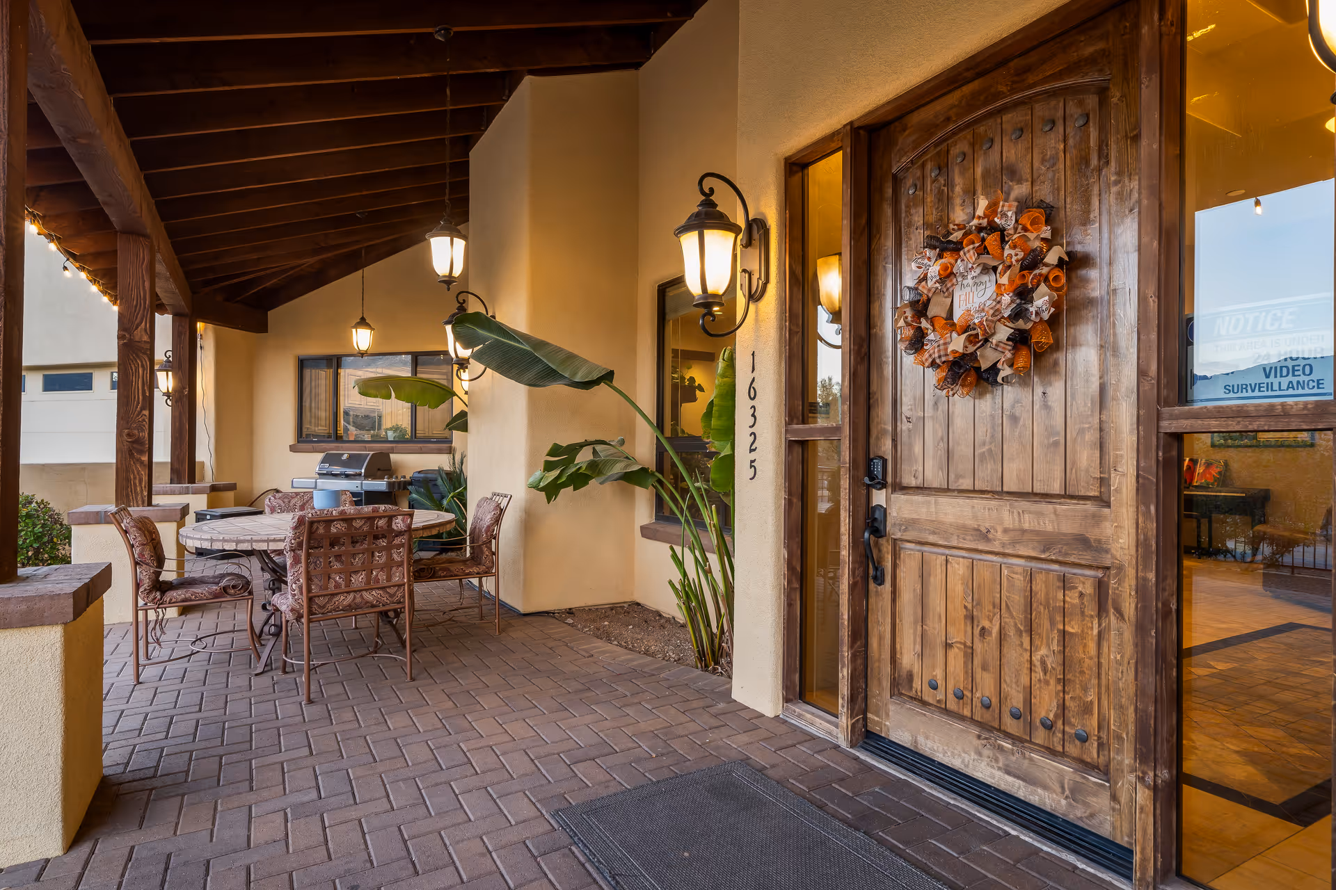 Covered outdoor patio area with a wooden front door decorated with a fall-themed wreath. The patio has a round table with four cushioned chairs, a barbecue grill, potted plants, and hanging lantern-style lights under a wooden roof.