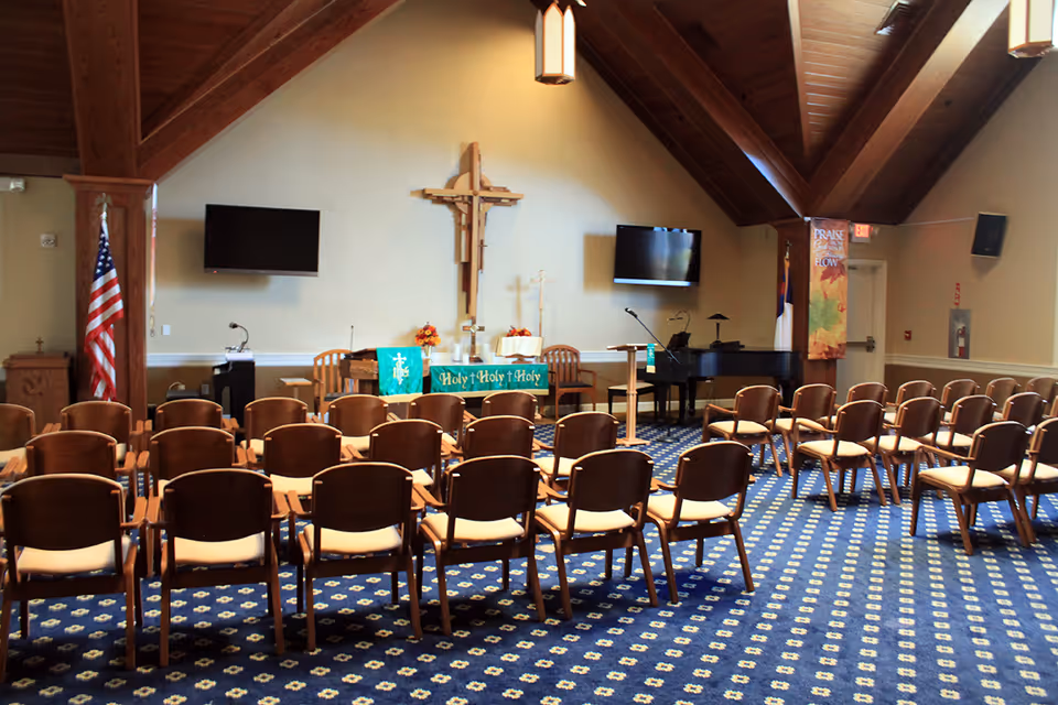 Interior of a chapel or worship room with rows of wooden chairs facing an altar adorned with green cloths that say 'Holy Holy Holy'. A large wooden cross is mounted on the wall behind the altar, flanked by two flat-screen TVs. The room has a high wooden ceiling with exposed beams, an American flag on the left, a piano on the right, and a carpet with a blue and yellow pattern.