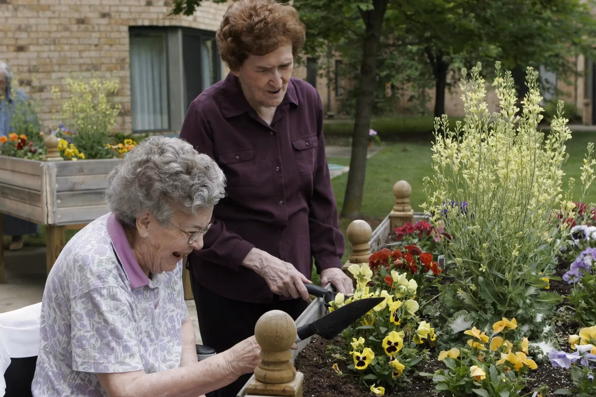 Two elderly women gardening together in an outdoor raised flower bed filled with colorful flowers and plants. One woman is seated and smiling while holding a gardening tool, and the other woman is standing beside her, also holding a gardening tool. The background shows a brick building and green trees.