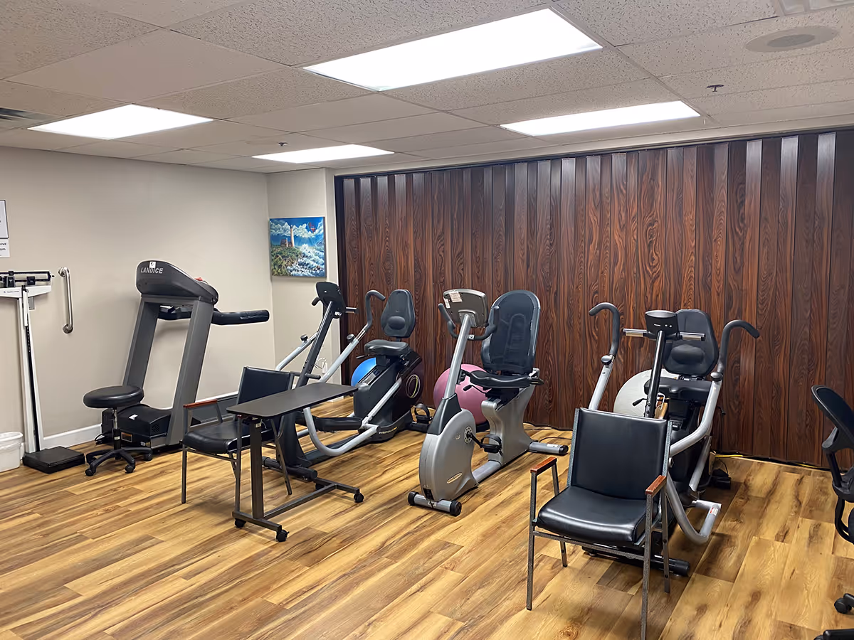 Indoor exercise room with a treadmill, multiple recumbent bikes, chairs and wood-look flooring against a wood-paneled wall.