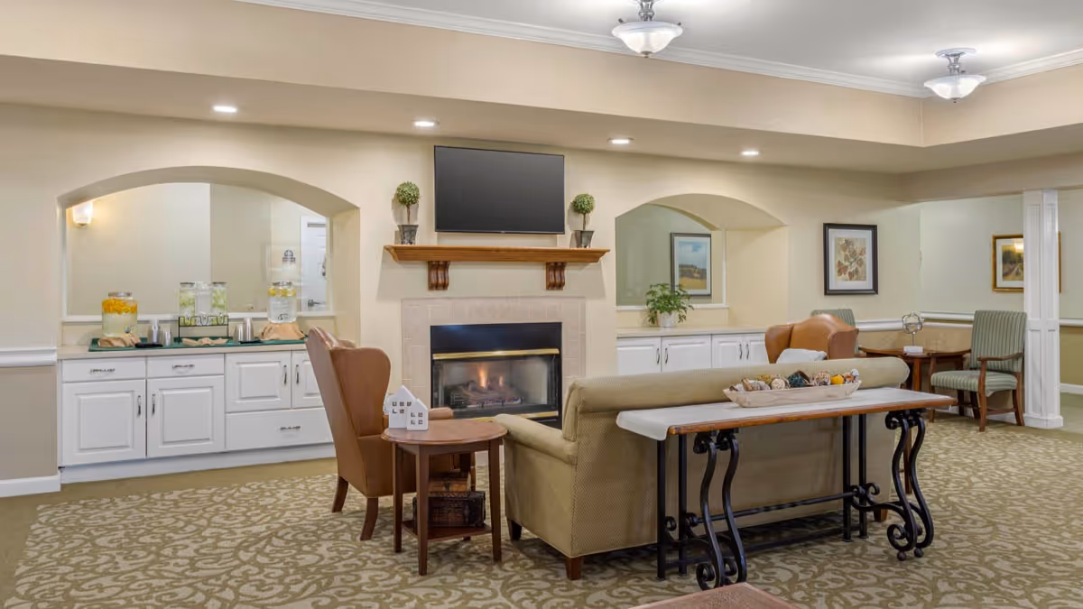 Comfortable senior living common room with sofas and chairs arranged around a fireplace and a refreshment buffet.