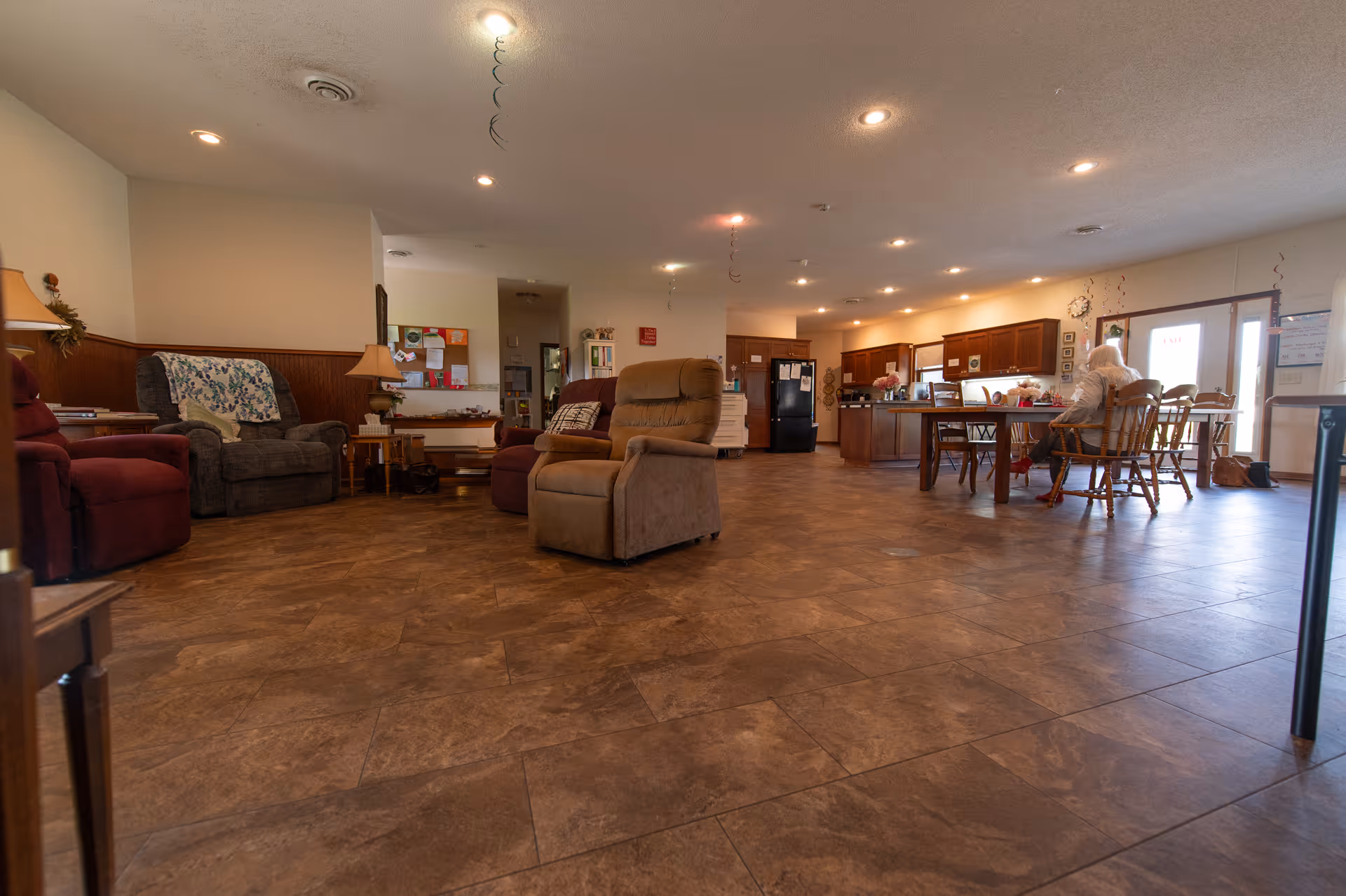 Spacious communal living area with recliners, a dining table near a kitchenette, and a person seated at the table.